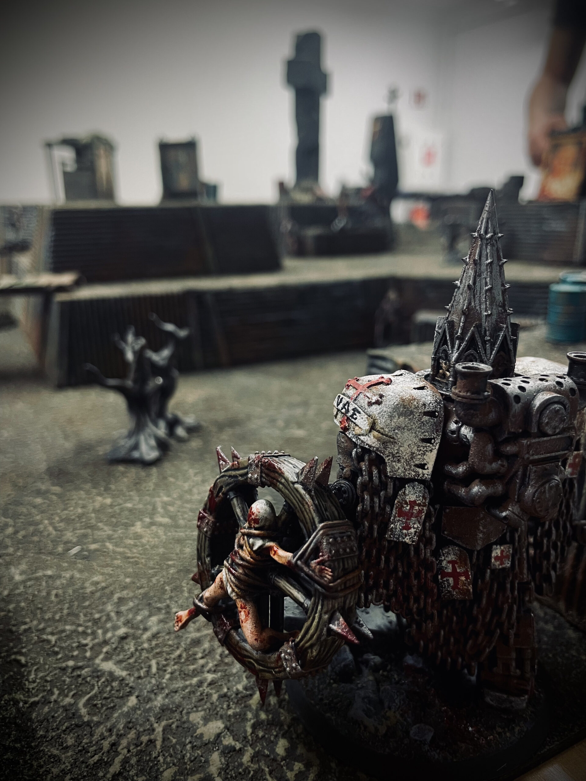 A cinematic over-the-shoulder view of a 28mm miniature looking across a Trench Crusade battlefield with HRC trenches, the Word of God pulpit, and the Concrete Cross in the distance.